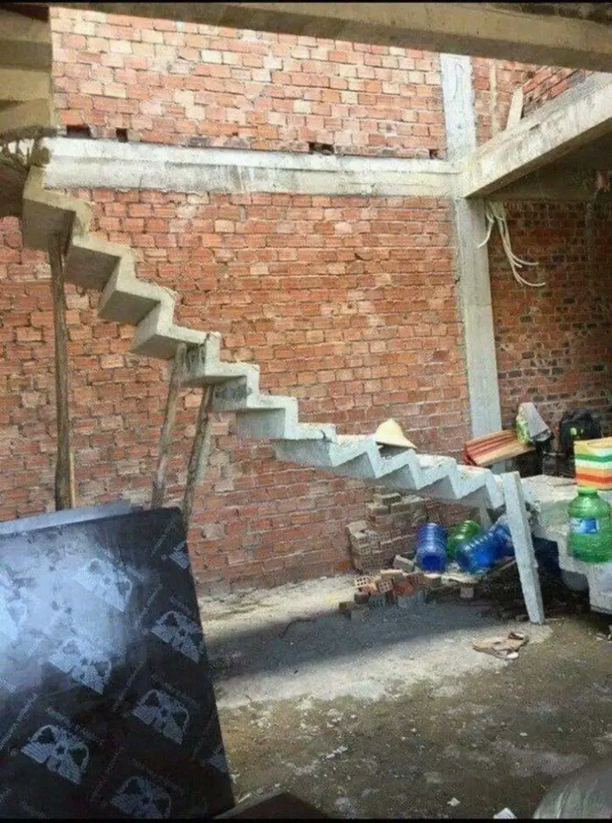 Unfinished concrete stairs without railings lead up along a rough brick wall in a construction site. Various building materials and plastic containers are scattered on the floor below.