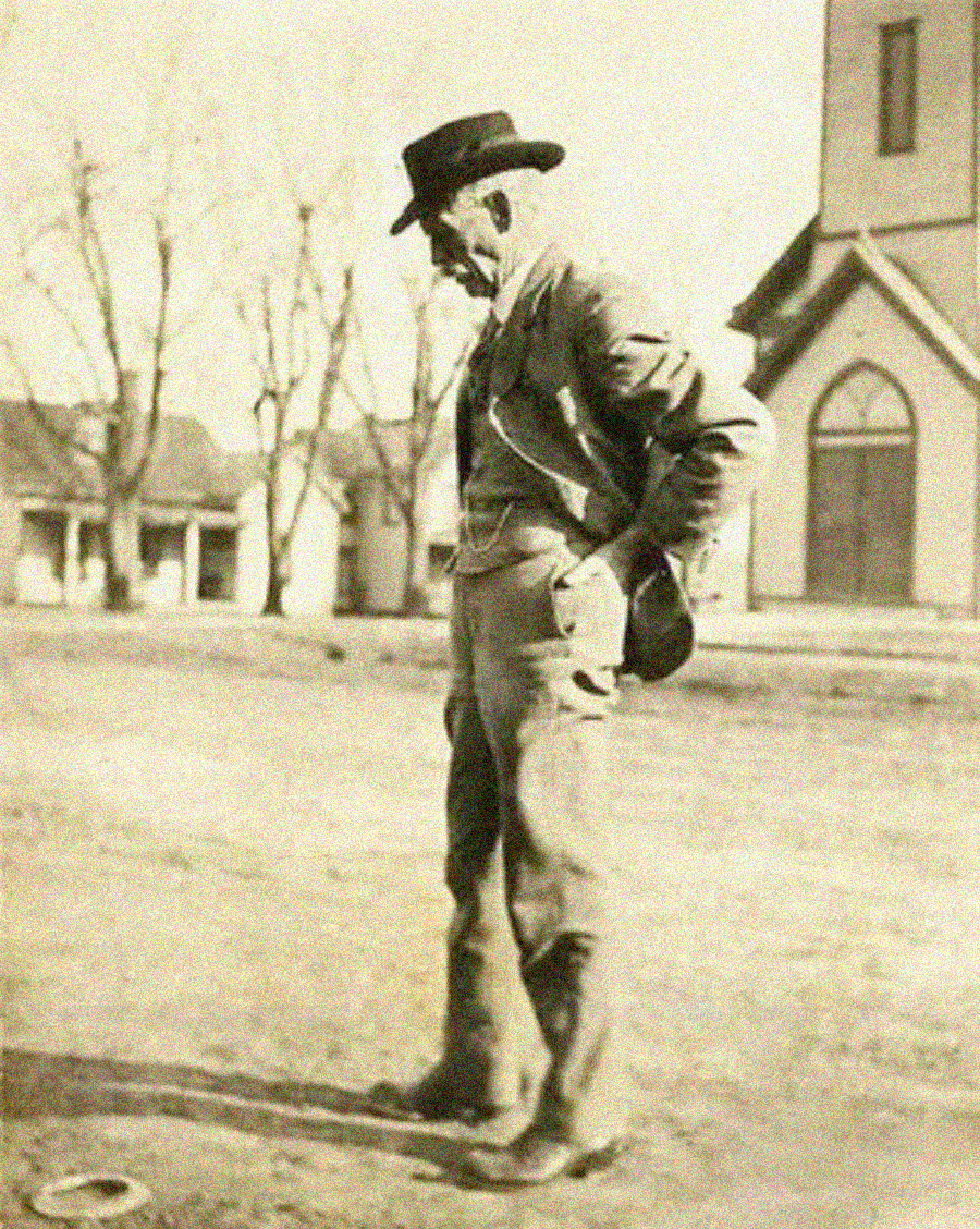 An elderly man in a hat and jacket stands outdoors on bare ground, looking down with his hands behind his back. Leafless trees and a building with arched windows are visible in the background.