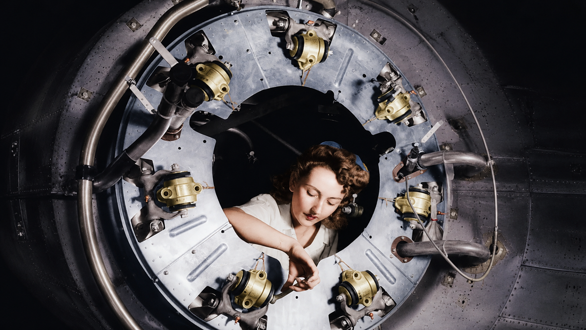 A woman works inside the circular opening of a large metal aircraft engine, surrounded by mechanical parts and gears, focused on her task.