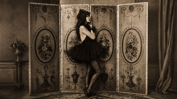 A woman in a black tutu and dark tights poses in front of an ornate, vintage folding screen with floral designs. The sepia-toned room has elegant decor and a vase of flowers on a side table. 1928