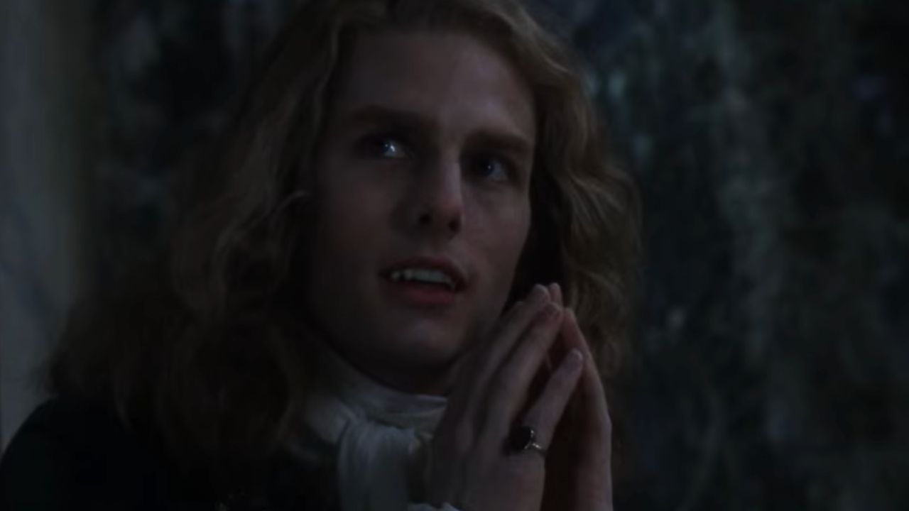 A person with long blond hair and pale skin clasps their hands together, gazing upward. They wear a dark coat and a white cravat, with dim lighting and a shadowy background.