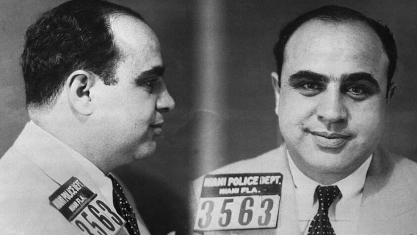 A black-and-white mugshot showing a man in a suit and tie from both the front and side, holding a placard labeled “Miami Police Dept. 3563” against a plain background.
