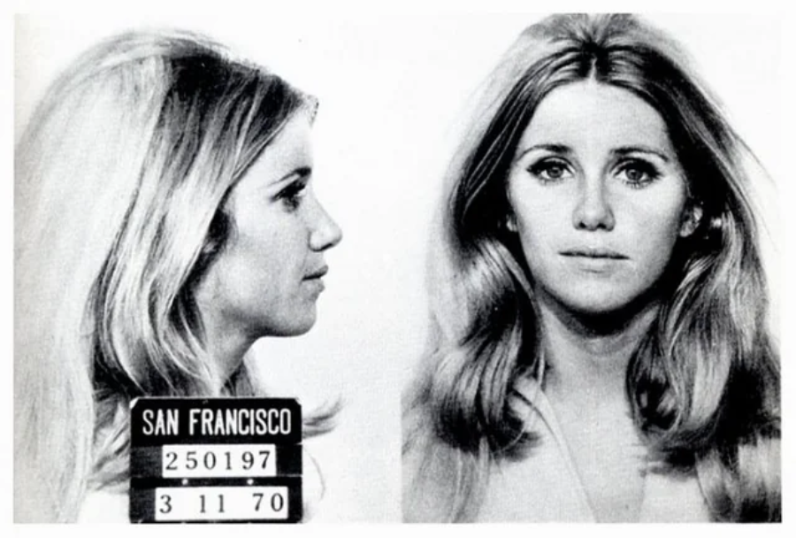Black-and-white mugshot of a woman with long light hair, shown in both profile and frontal views. A sign reads "San Francisco" with booking numbers and the date 3-11-70.