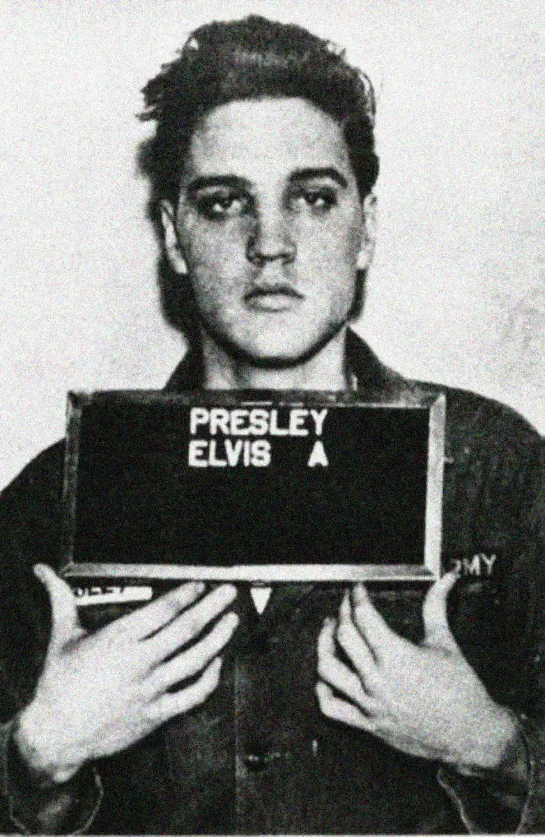 A young man holds a nameplate reading "PRESLEY ELVIS" in front of his chest, facing the camera for a black-and-white mugshot-style photograph. He wears a military-style shirt.