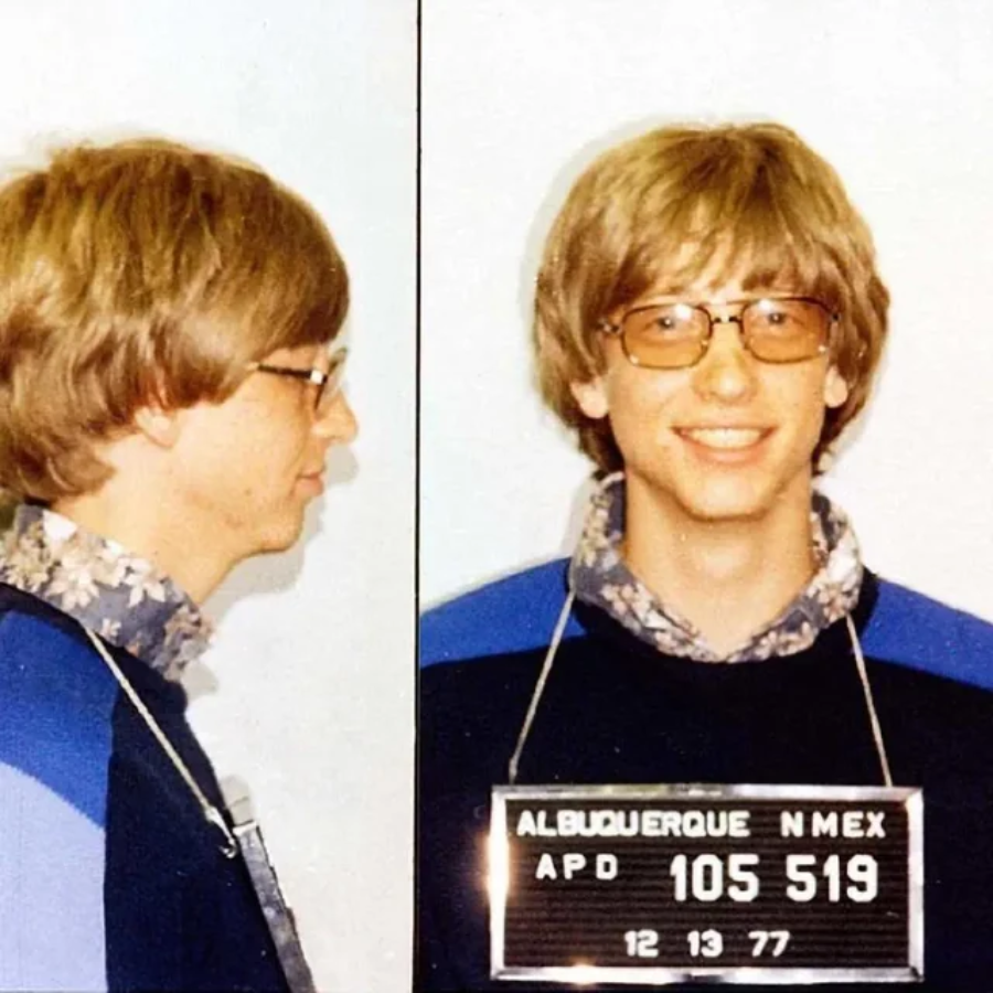 A young man with blond hair and glasses is shown in a mugshot, smiling. He wears a floral shirt under a dark sweater. The mugshot plaque reads "Albuquerque N. Mex APD 105519 12 13 77.