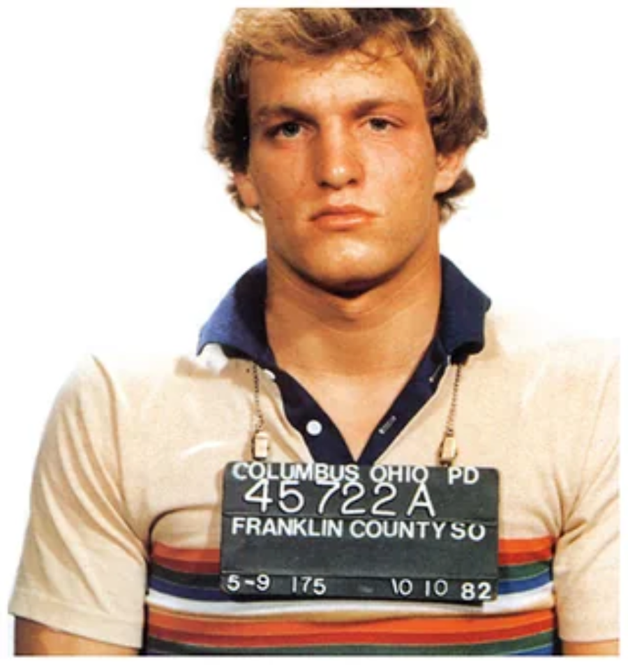 Young man with short blond hair wearing a striped polo shirt, holding a sign with arrest details from Columbus, Ohio Police Department, Franklin County, dated 10/10/82.