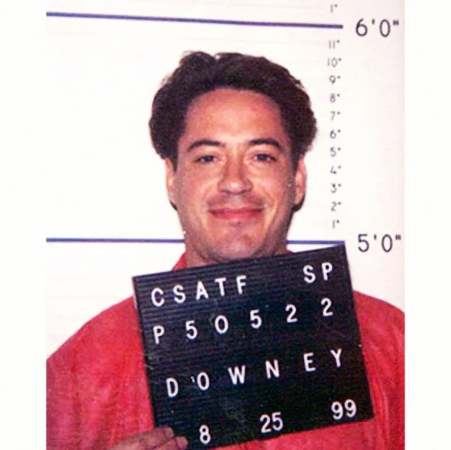 A man in a red shirt stands against a height chart, smiling and holding a black sign with white letters displaying “CSATF SP P50522 DOWNEY” and a date “8 25 99.”