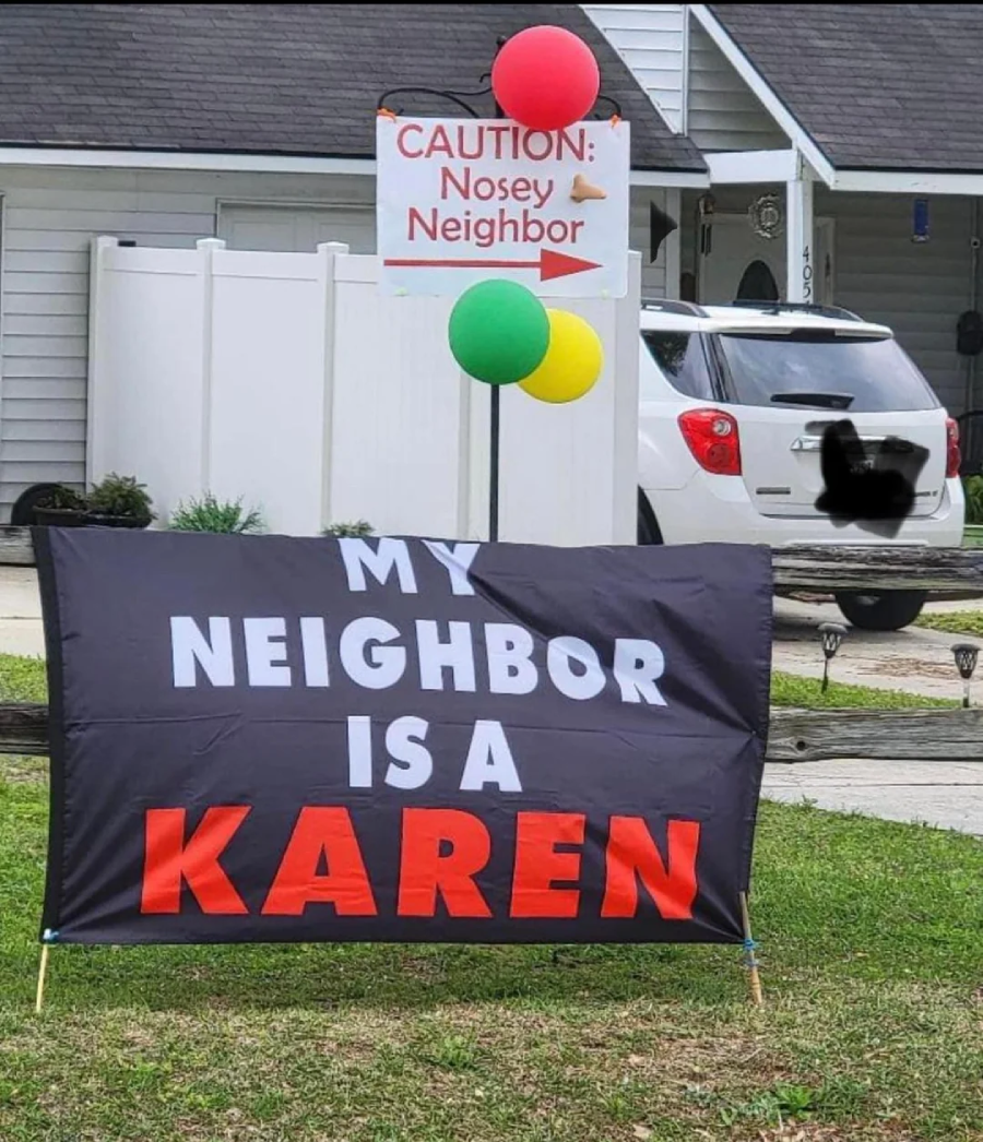A yard sign reads “MY NEIGHBOR IS A KAREN” in bold letters. Behind it, a sign with balloons says “CAUTION: Nosey Neighbor” with a pointing hand, in front of a white house and parked car.