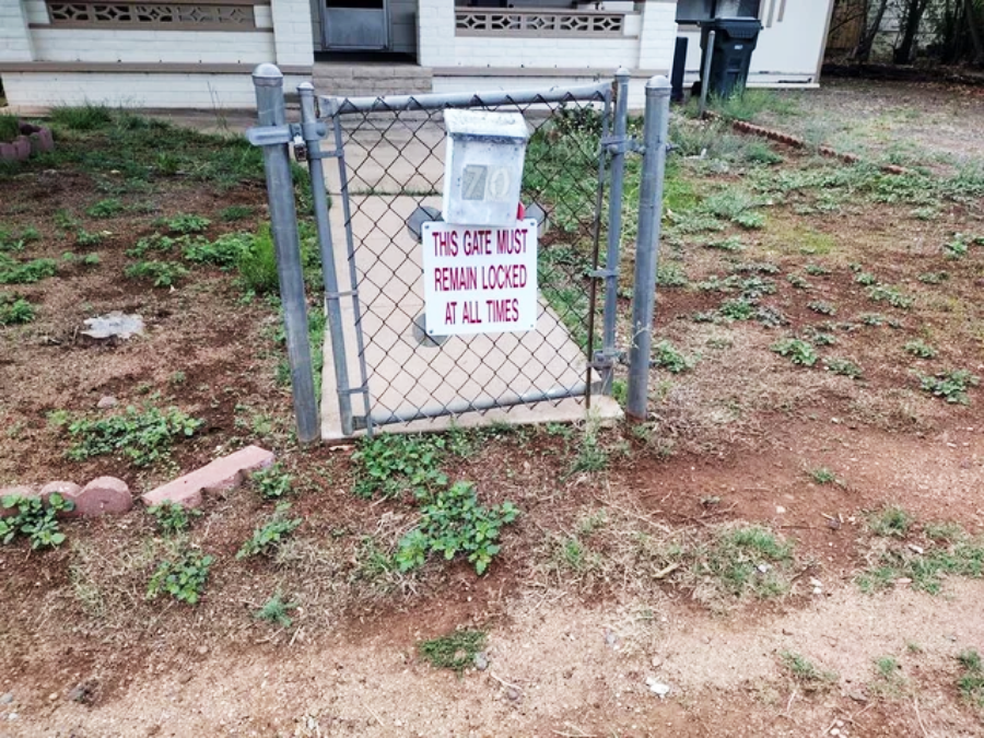 A chain-link gate with a sign that reads "THIS GATE MUST REMAIN LOCKED AT ALL TIMES," but the gate is unlocked and open. There is a house and patchy grass in the background.