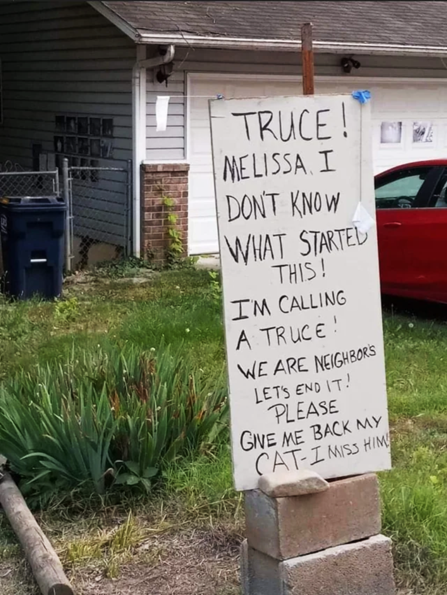 A handwritten sign in a yard reads: “Truce! Melissa, I don’t know what started this! I’m calling a truce! We are neighbors, let’s end it! Please give me back my cat. I miss him.” A house and a red car are in the background.
