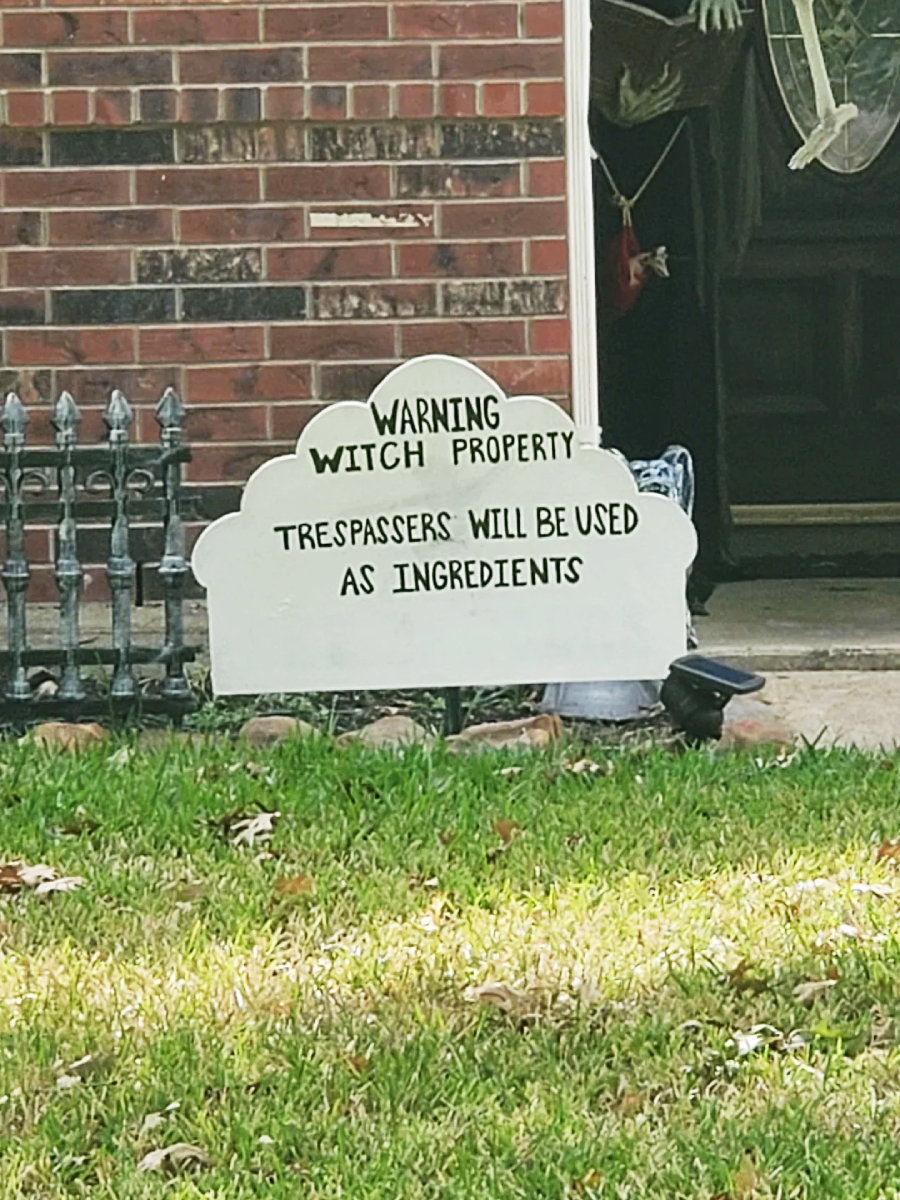 A white sign on a lawn reads, "Warning Witch Property. Trespassers will be used as ingredients." It sits in front of a brick house with Halloween decorations, including a fake boot and fence.