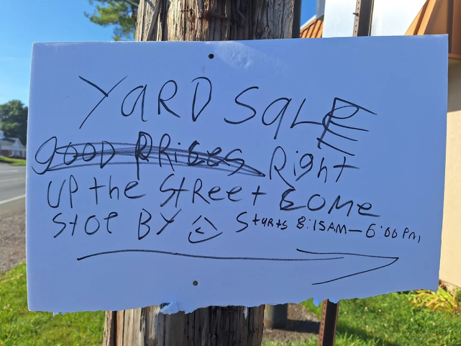 A handwritten sign on a pole reads: "YARD SALE. Good prices right up the street & come stop by :), starts 8:15am–6:00pm," with a right-pointing arrow. Some words are crossed out.