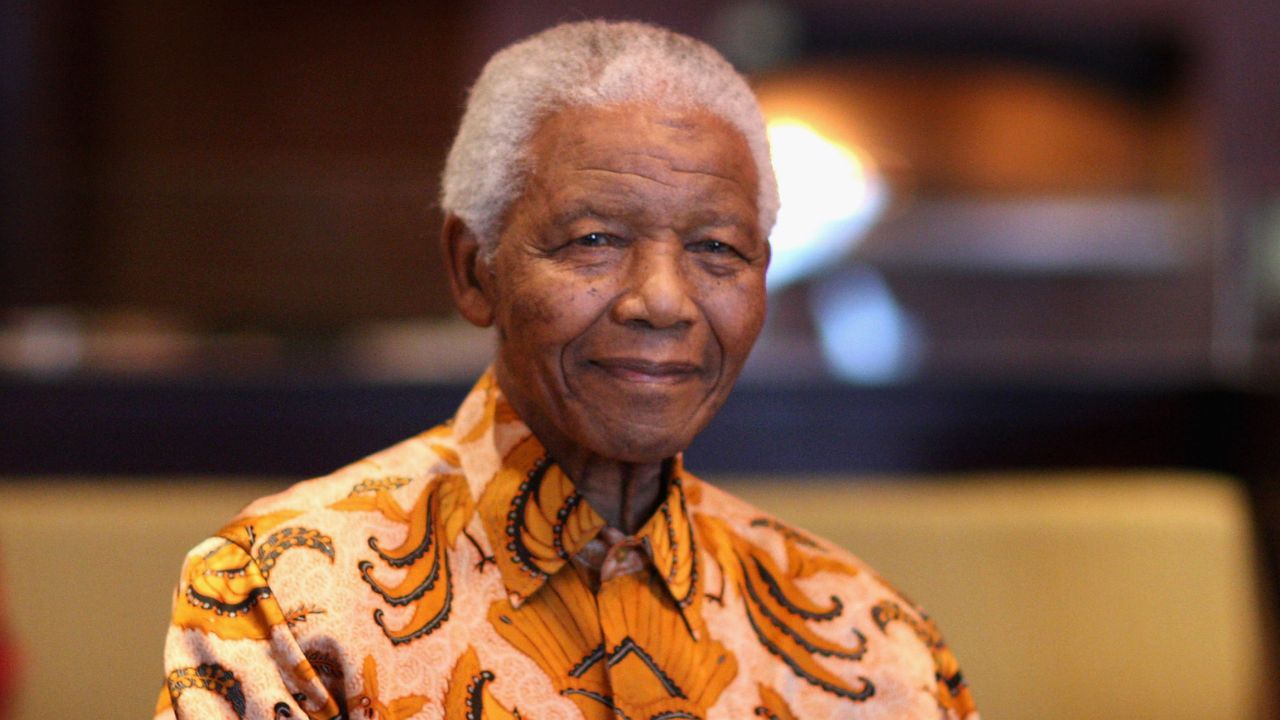 An elderly man with gray hair smiles gently while wearing a bright patterned shirt, seated indoors with a blurred background.