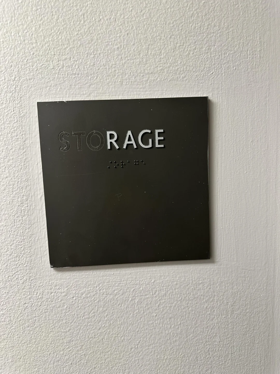 A black sign on a white wall reads "STORAGE" in raised silver letters, with Braille text underneath. The lighting casts slight shadows on the wall.