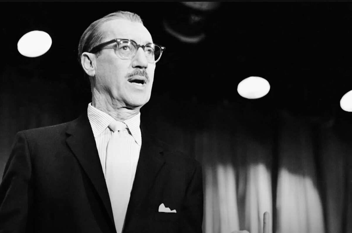 A man in a suit, tie, and glasses stands under stage lights, looking to the side with a serious expression. There are curtains in the background. The image is in black and white.