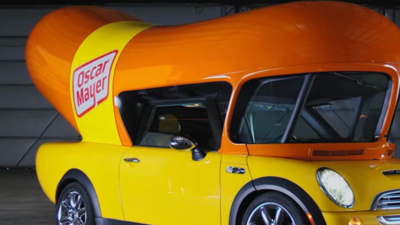 A yellow car with a large orange hot dog-shaped structure on top, featuring the Oscar Mayer logo, is parked indoors.
