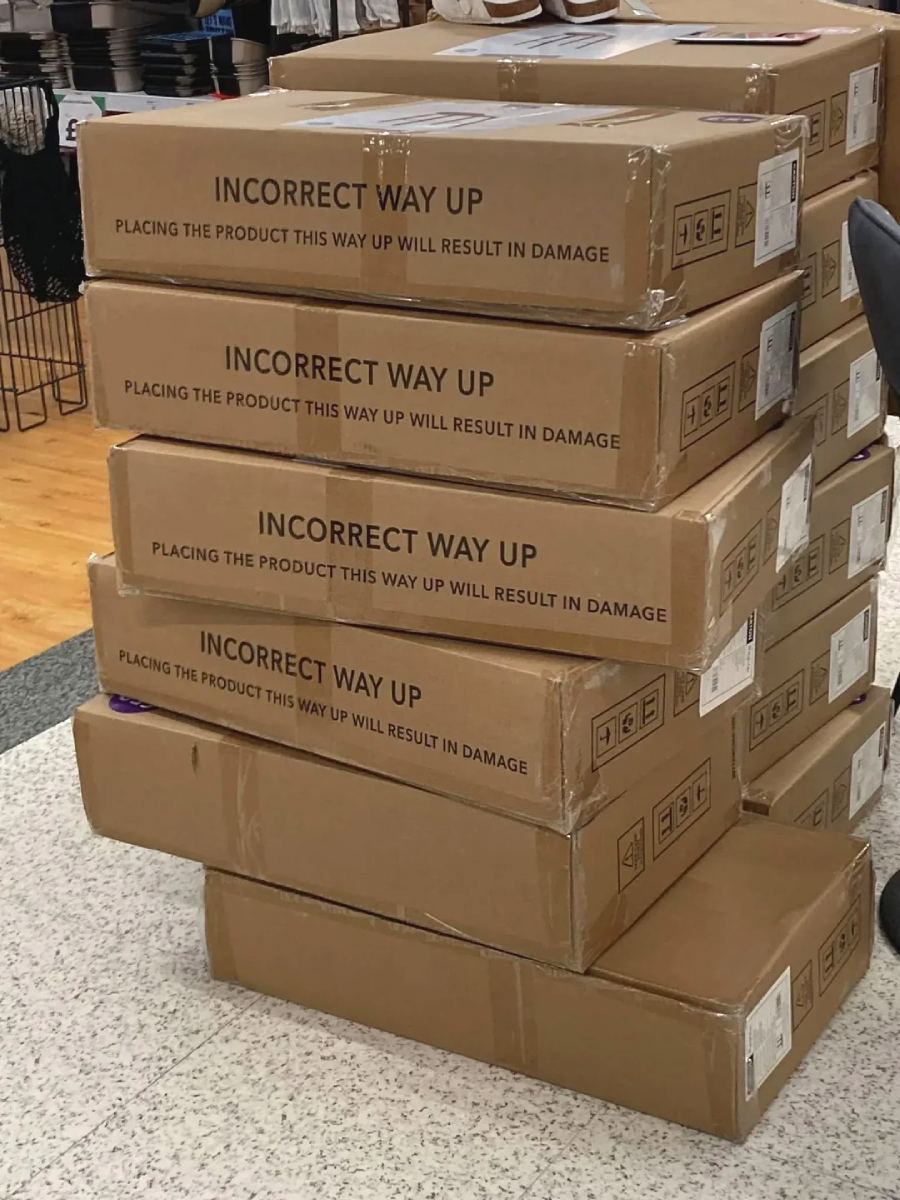 Six cardboard boxes are stacked on a counter. Each box is labeled with the text “INCORRECT WAY UP. PLACING THE PRODUCT THIS WAY UP WILL RESULT IN DAMAGE,” though all the boxes are stacked in that incorrect orientation.