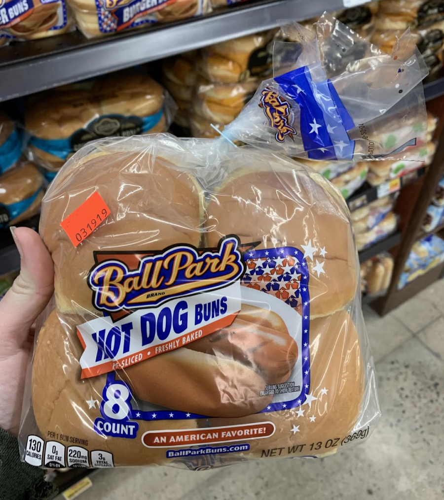 A hand holding a package of Ball Park brand hot dog buns in a grocery store. The package contains 8 sliced buns and has a price sticker reading $3.19. Other bread products are visible on the shelf behind.