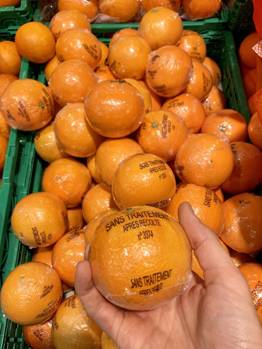 A hand holds an orange wrapped in plastic with French text on it, above a pile of similarly wrapped oranges displayed in a green crate at a market or grocery store.