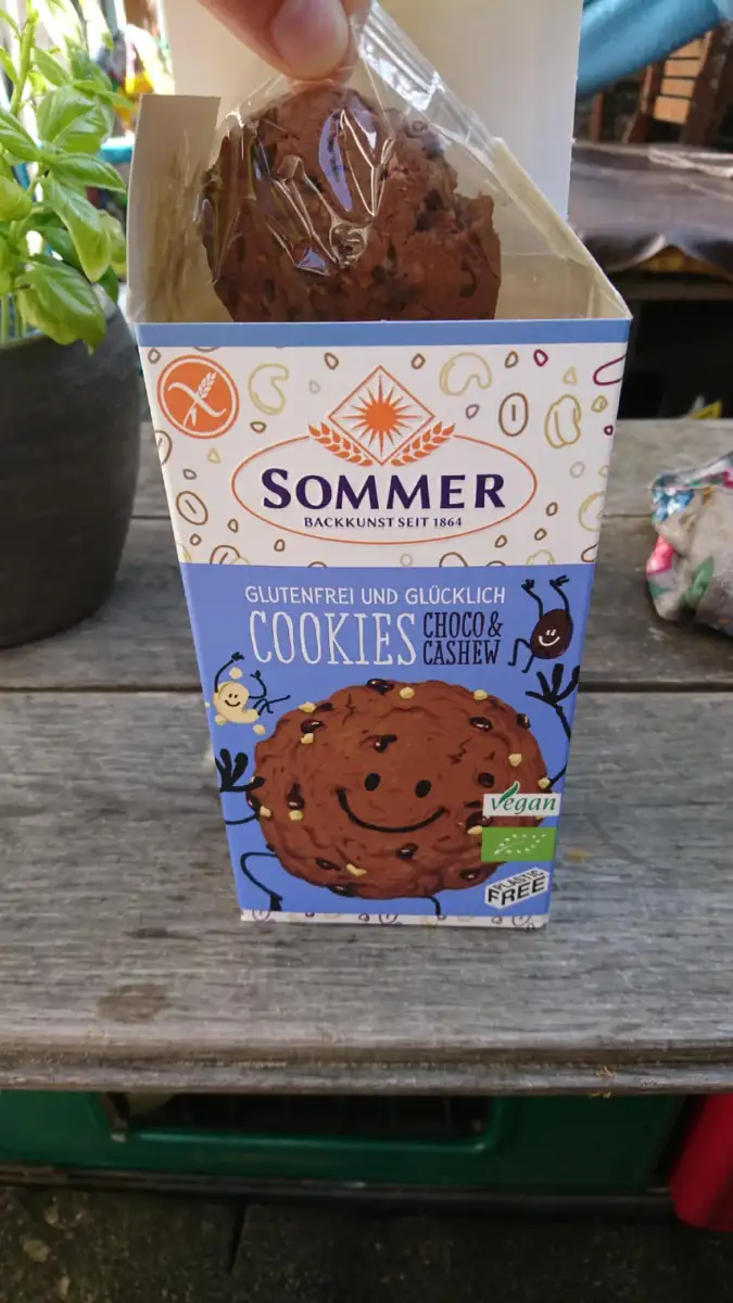 A hand holds an open box of Sommer brand gluten-free and vegan choco & cashew cookies on a wooden table, with a potted plant and colorful fabric in the background.