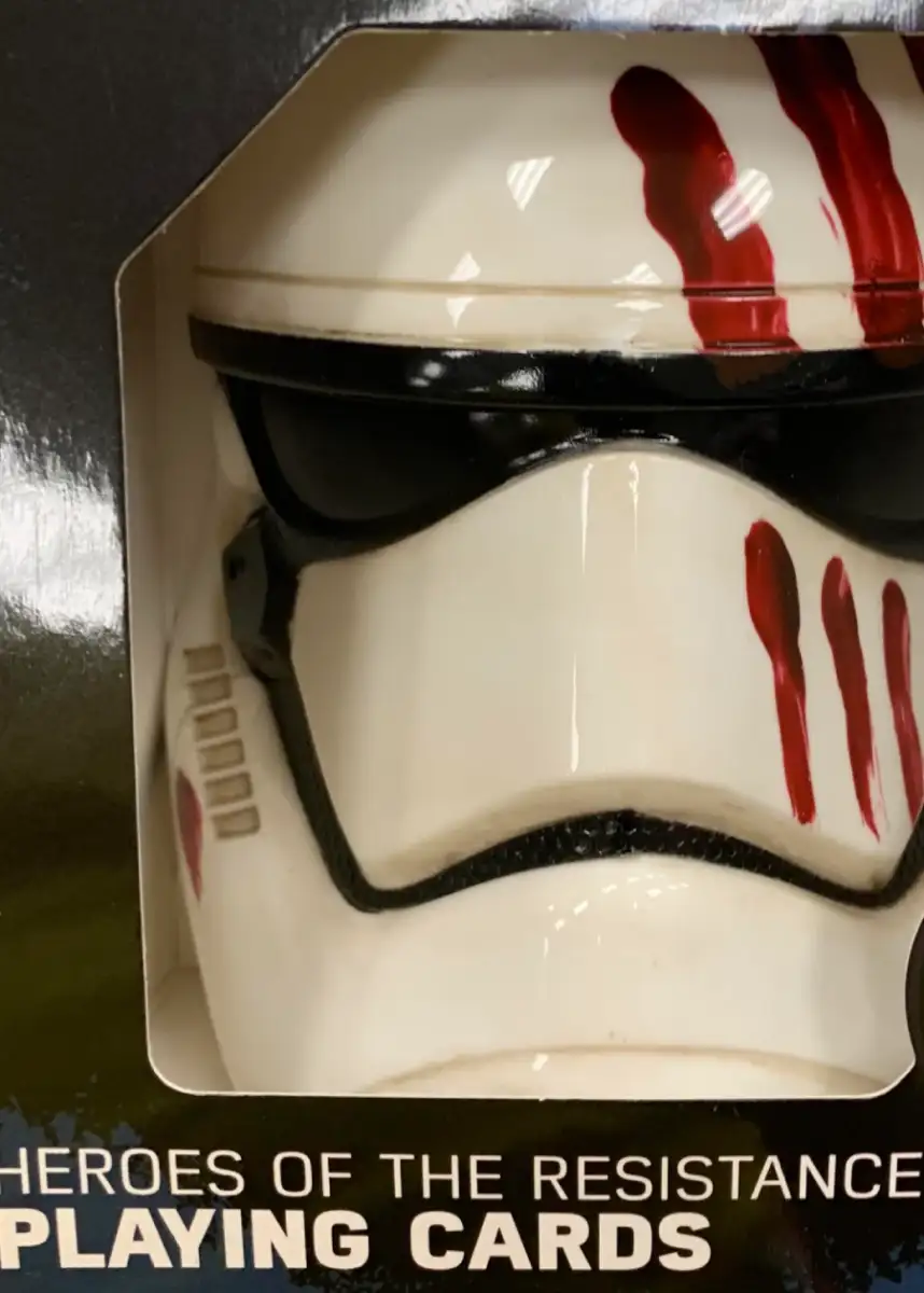 A close-up of a Star Wars Stormtrooper helmet toy with red paint streaks across the top and right side, in packaging labeled "HEROES OF THE RESISTANCE PLAYING CARDS.
