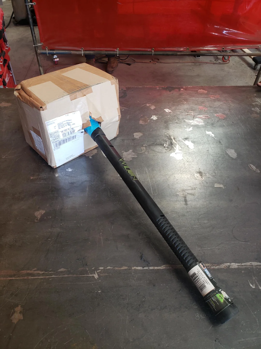 A large black metal rod is sticking through a cardboard shipping box on a concrete floor in an industrial setting. The box is slightly crushed where the rod penetrates it.