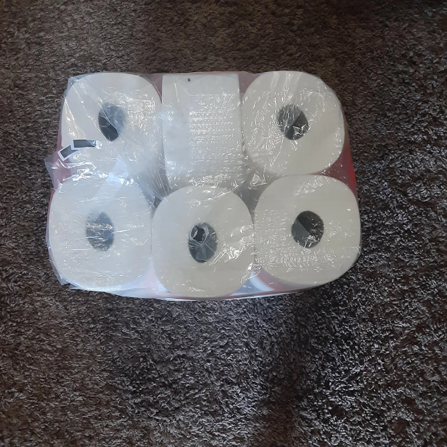 A six-pack of white toilet paper rolls wrapped in clear plastic sits on a brown textured carpet.