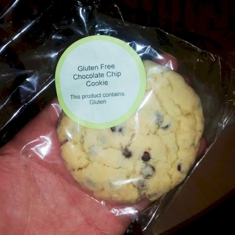 A hand holds a packaged cookie labeled "Gluten Free Chocolate Chip Cookie." The label also states, "This product contains: Gluten," creating a contradiction.