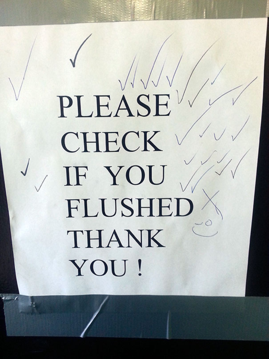 A paper sign reads "PLEASE CHECK IF YOU FLUSHED THANK YOU!" in bold text, covered with multiple hand-drawn check marks and a small smiley face. The sign is taped to a surface.