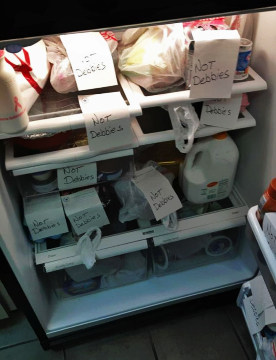 A refrigerator filled with various food items, each labeled with handwritten notes saying “Not Debbie’s” on paper taped to the items, indicating they belong to someone other than Debbie.