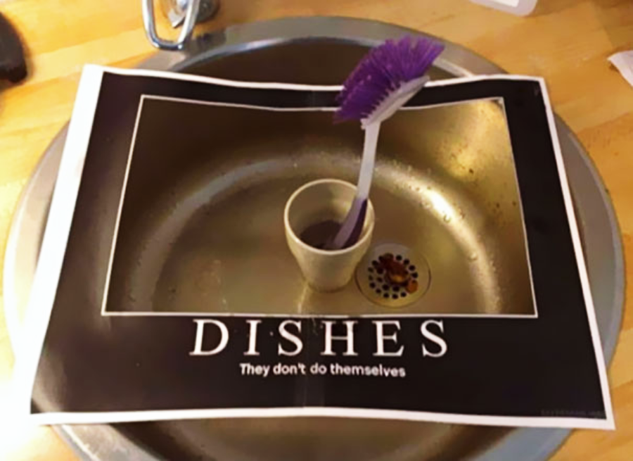 A kitchen sink with a motivational-style poster that says “DISHES: They don’t do themselves.” A cleaning brush in a cup is placed on top of the poster inside the sink.