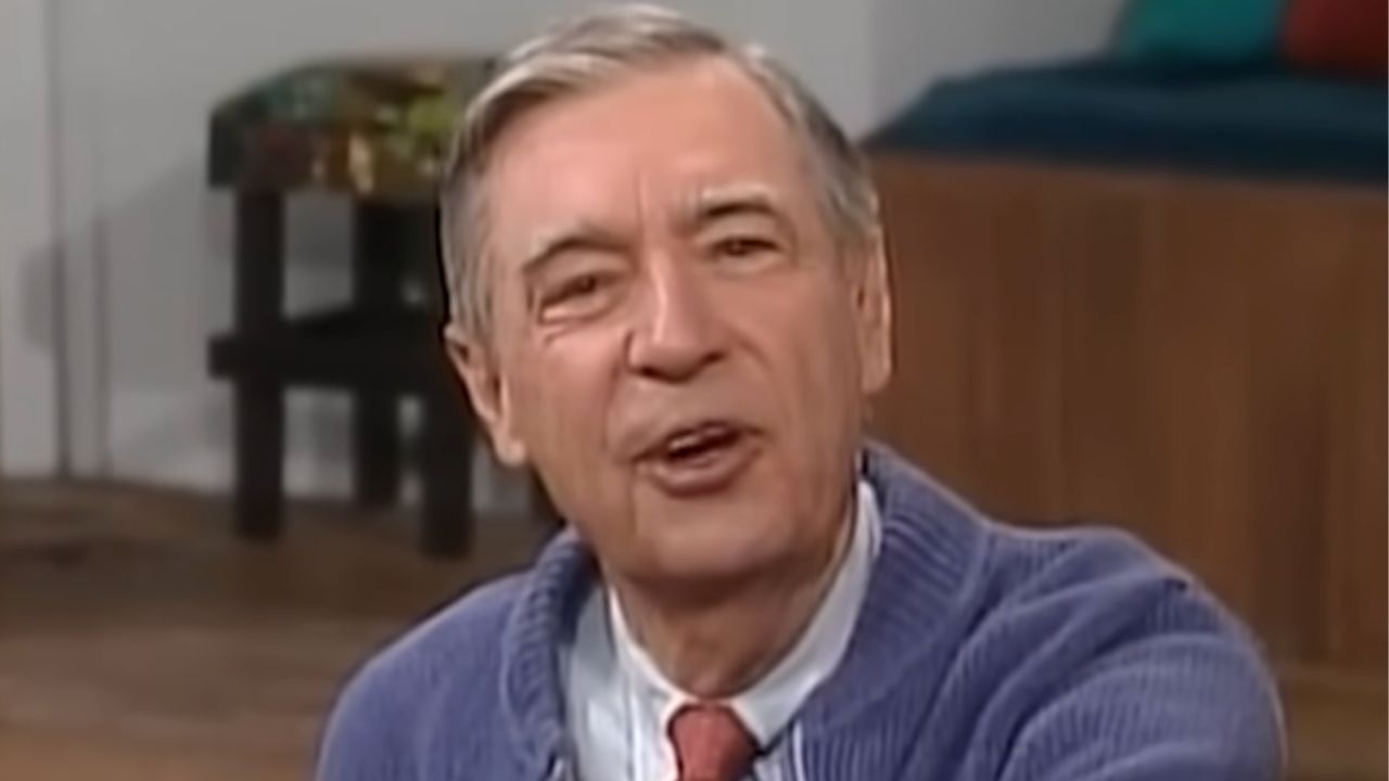 An older man with gray hair, wearing a blue sweater and red tie, speaks while seated indoors. The background includes a wooden bench, colorful pillows, and part of a small table.