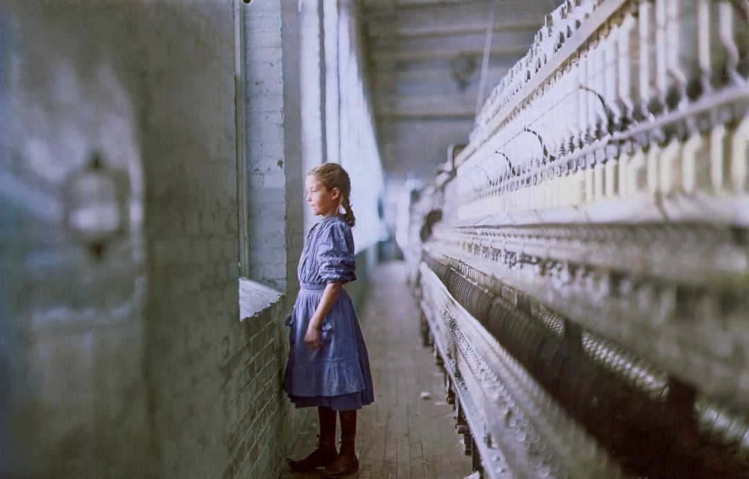 A young girl in a blue dress stands by a window in a cotton mill, looking outside. She is surrounded by large spinning machines in a long, narrow, industrial room.