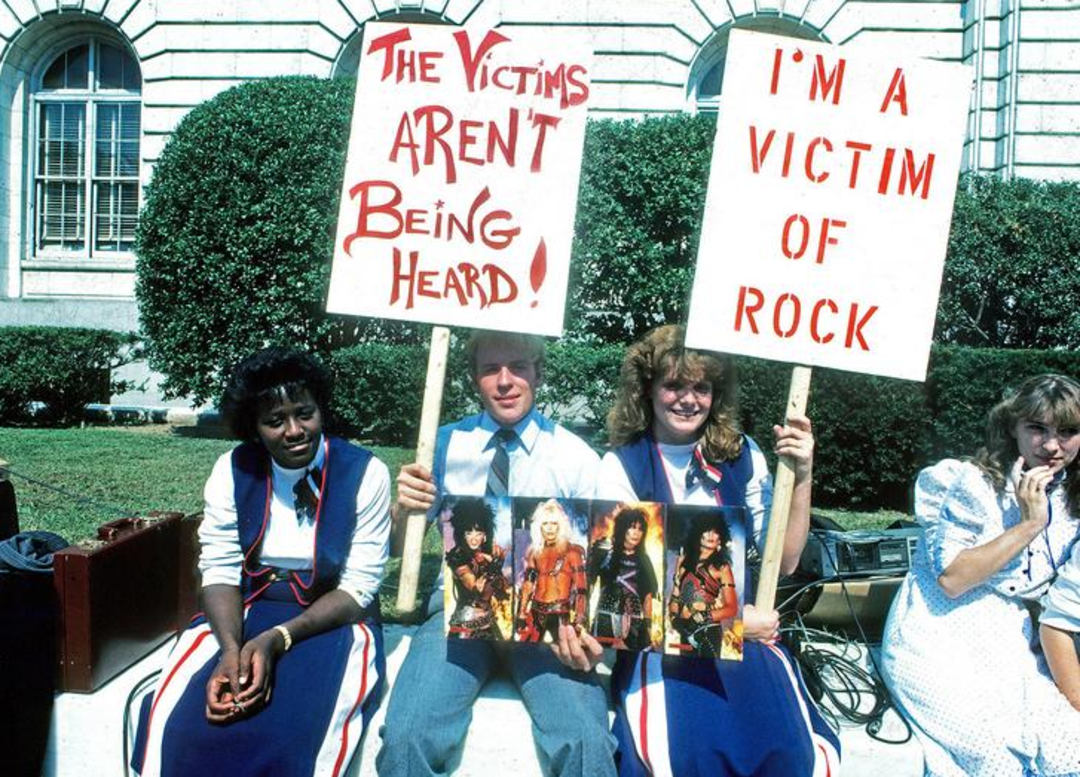 Three young people sit outside on a curb. Two hold protest signs; one reads "The victims aren't being heard!" and the other says "I'm a victim of rock." They display photos of a glam rock band.