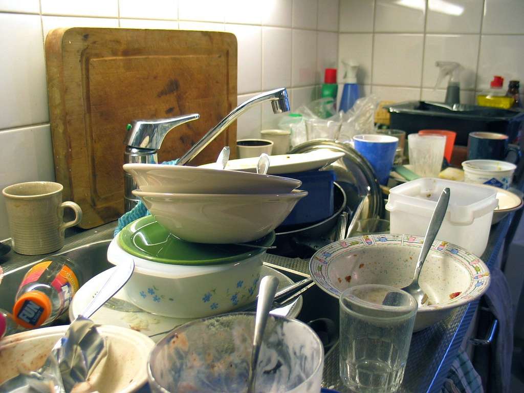 A kitchen sink overflowing with dirty dishes, bowls, cups, and utensils stacked beside and inside the sink; cleaning supplies and a cutting board are visible in the background.