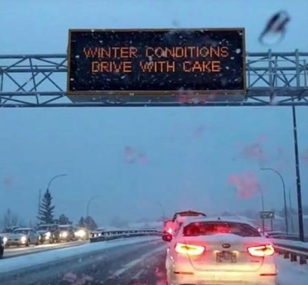 A snowy road with cars lined up and a highway sign overhead that reads, "WINTER CONDITIONS DRIVE WITH CAKE" instead of "care.