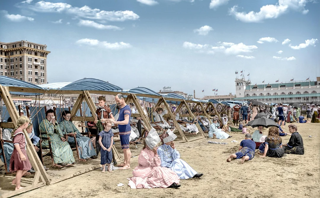 People in vintage clothing relax under shaded wooden structures on a crowded sandy beach. Some are seated, others stand or sit on the sand, while buildings and blue sky with clouds are visible in the background.