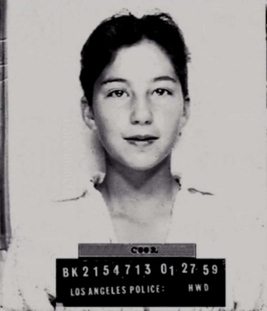 Black and white mugshot of a young woman with dark hair, wearing a light-colored top, looking at the camera. The placard reads "BK2154713 01-27-59 LOS ANGELES POLICE: HWD.