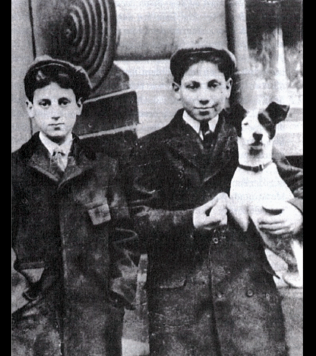 Two boys in coats and caps stand side by side; the boy on the right is holding a small dog. The photo is black and white and appears to be old, suggesting it was taken in the early 20th century.
