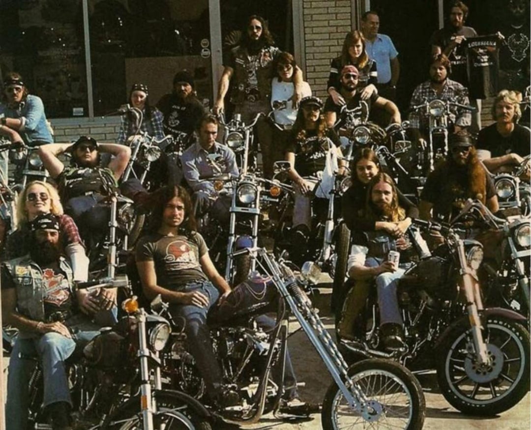 A large group of bikers, both men and women, sit on or stand near their motorcycles in front of a building, posing for a photo. Most wear sunglasses, jeans, and leather vests, exuding a rugged, laid-back attitude.