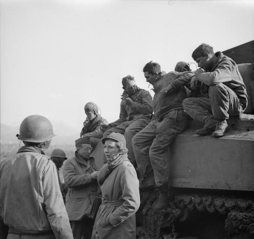 A group of soldiers and a woman in a coat stand and sit on a tank, talking and resting. Some soldiers wear helmets, while others sit casually on the tank’s side. The scene appears to be from a historical wartime setting.