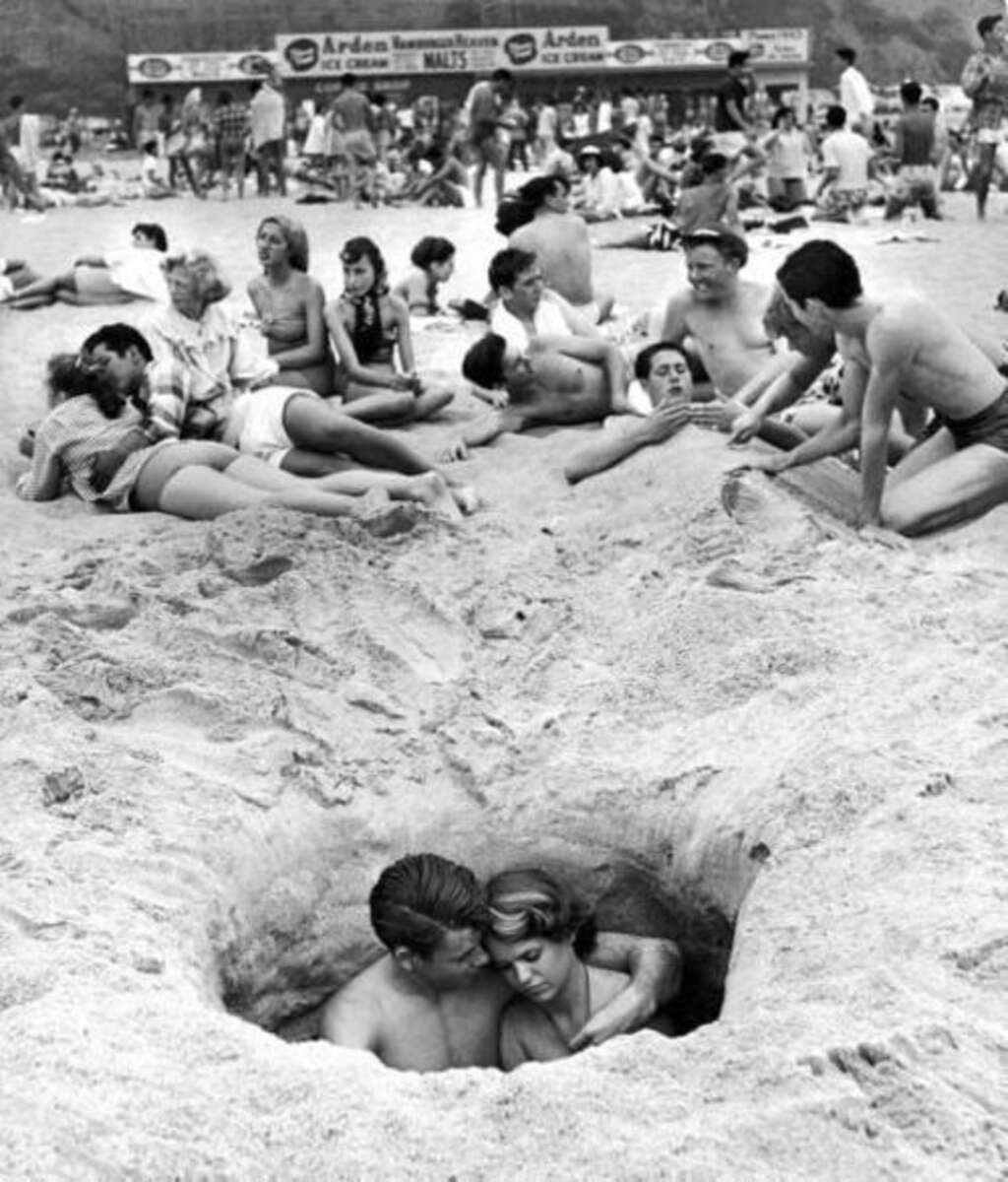 A young couple sits together in a large hole dug in the sand on a crowded beach, surrounded by groups of sunbathers and people relaxing near an ice cream stand in the background.
