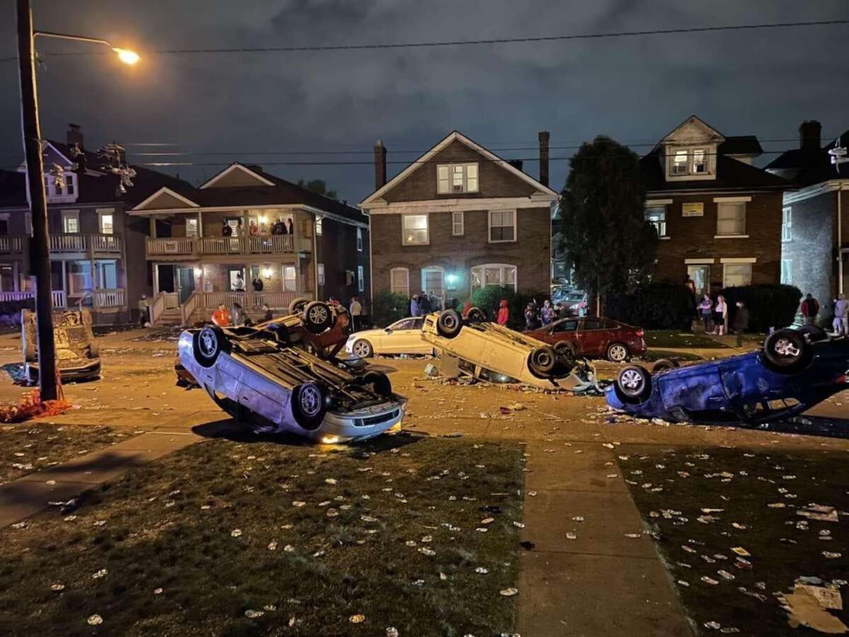 Several cars are overturned on a suburban street at night, with debris scattered across the road and lawns. People stand on sidewalks and porches in front of houses, observing the scene.