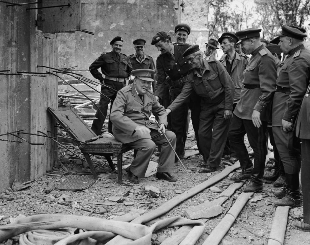 A group of uniformed men stand around a seated figure in military attire, surrounded by rubble and debris in a damaged building, appearing to converse and smile. Exposed rebar and pipes are visible in the scene.
