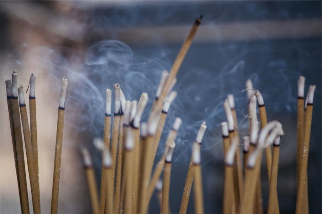 A cluster of burning incense sticks emits thin trails of smoke against a blurred background, creating a serene and atmospheric scene.