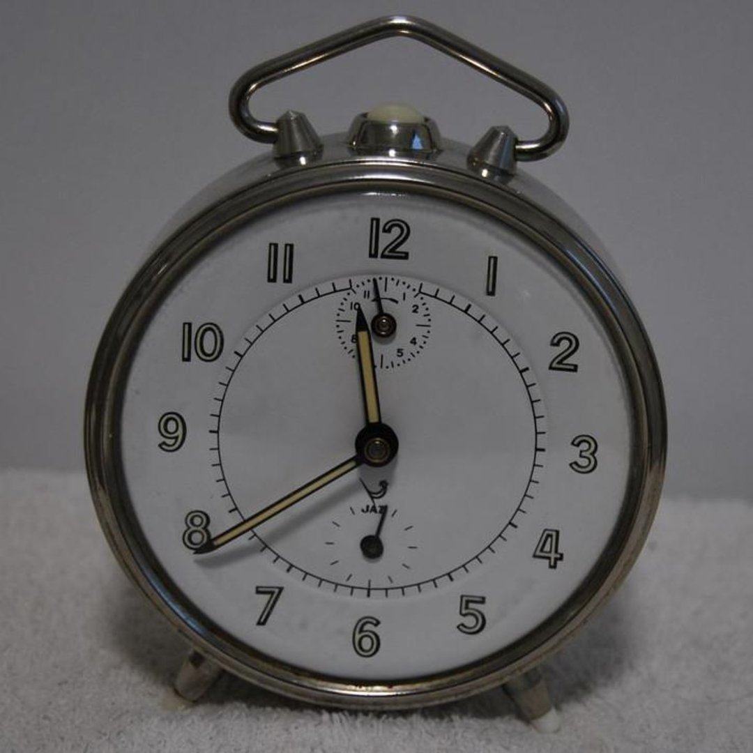 A vintage silver analog alarm clock with black numbers and hands sits on a white surface. The time on the clock shows 7:00. There is a smaller subdial above the center of the clock face.