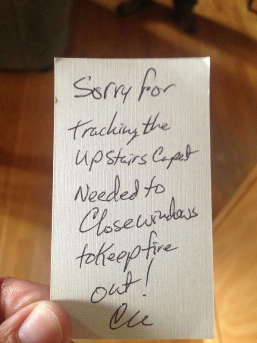 A handwritten note reads: "Sorry for tracking the upstairs carpet. Needed to close windows to keep fire out! Cie." A hand holds the note over a wooden floor.