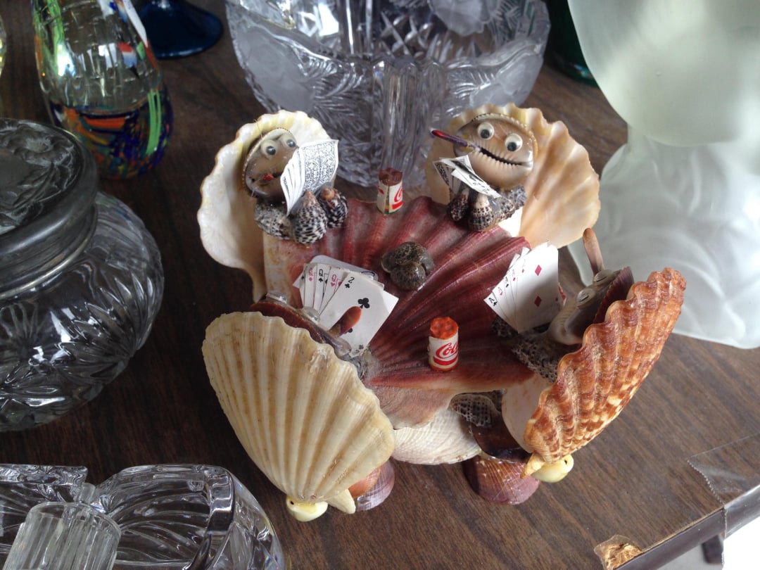 Three figurines made from seashells and other materials sit around a table, playing cards and drinking. Each figure is holding playing cards, with beer bottles and additional cards on the table.