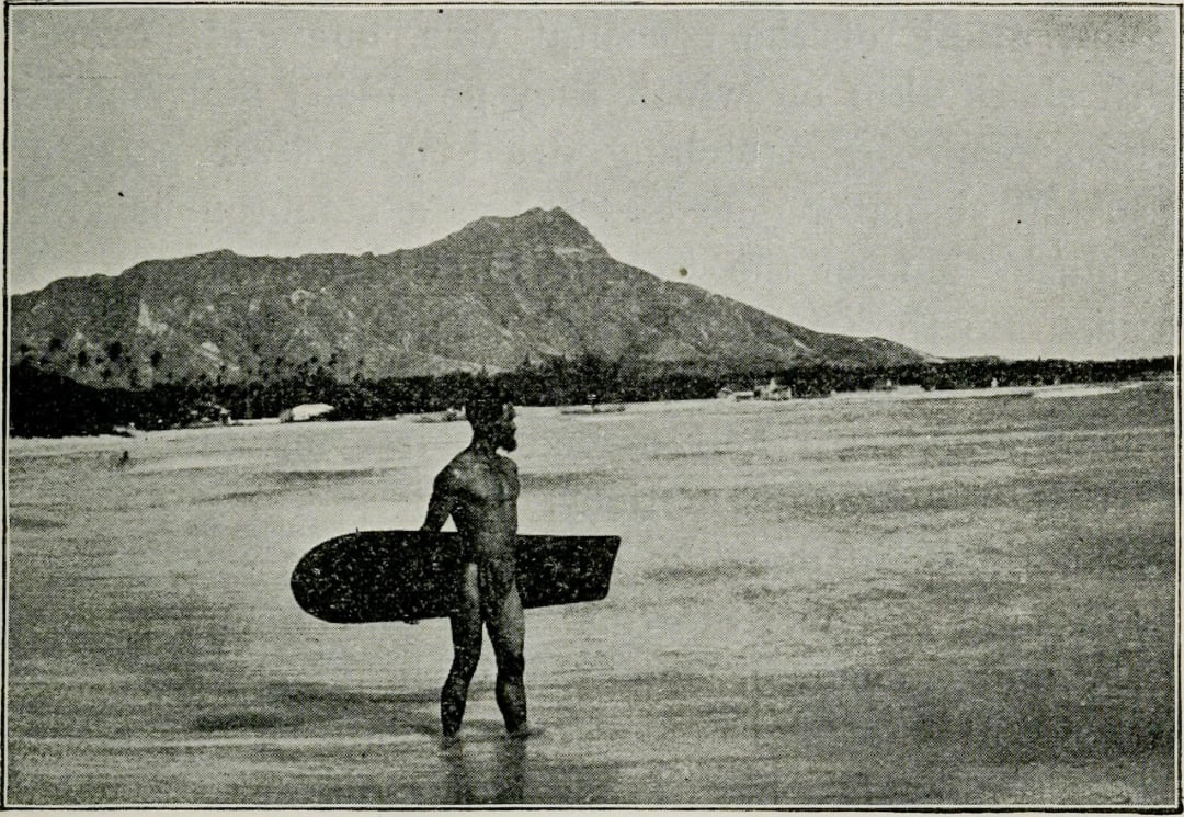 A person stands in shallow water holding a surfboard, with a large mountain in the background under a cloudy sky.