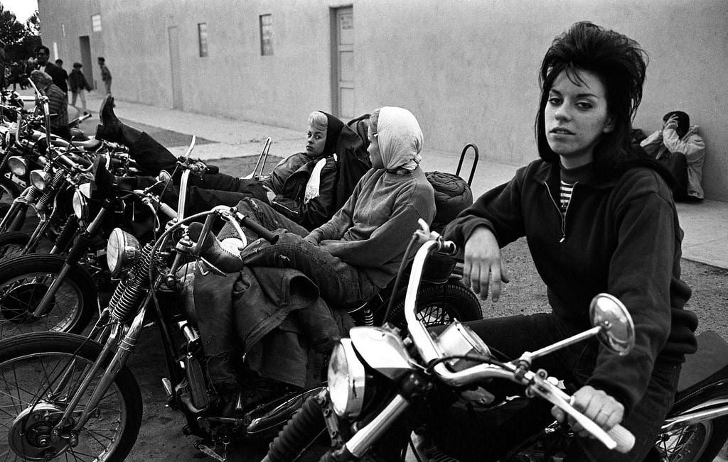 A woman with voluminous hair sits confidently on a motorcycle in the foreground, while others relax on nearby bikes in a row beside a plain building. Several people are visible in the background on the sidewalk.